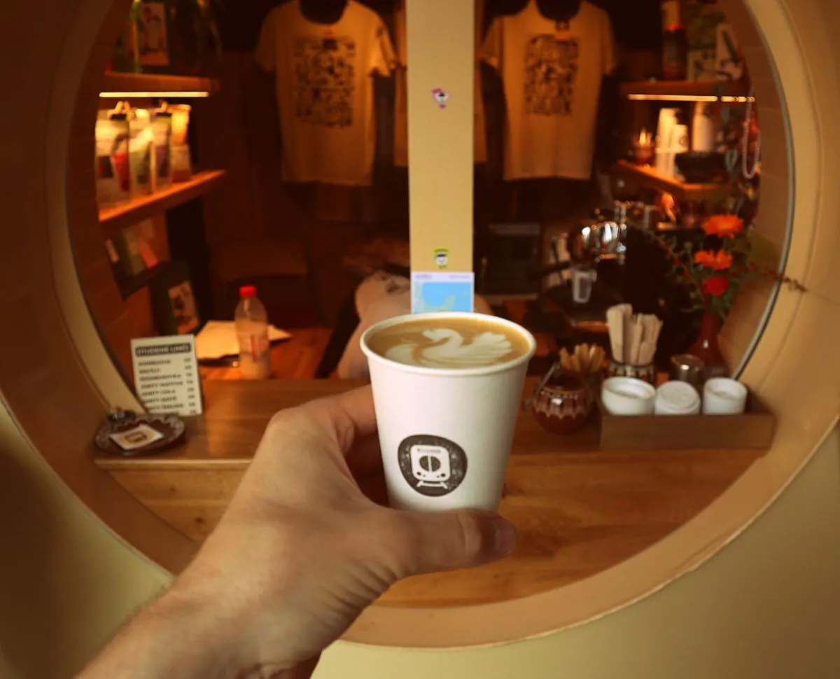 Ruka drží kelímek kávy Metro Kaffe s labutí latté art před okénkem